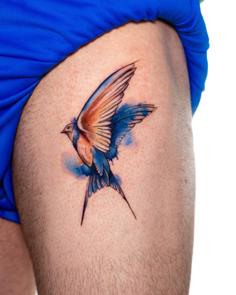 Watercolor Hummingbird Tattoo on Upper Thigh, with blue and orange watercolor splashes.