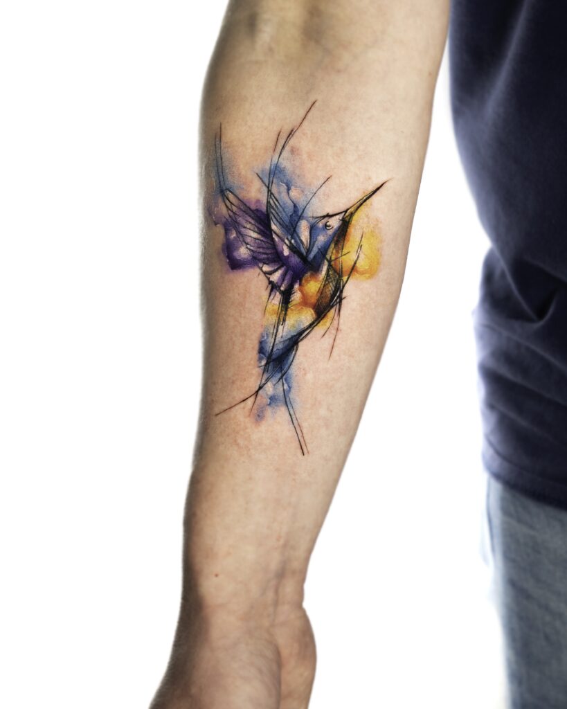 A Watercolor Hummingbird Tattoo on arm, with blue, purple and orange watercolor splashes.